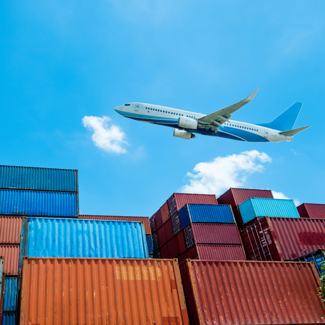 A large cargo airplane flies high above a stack of colorful shipping containers.